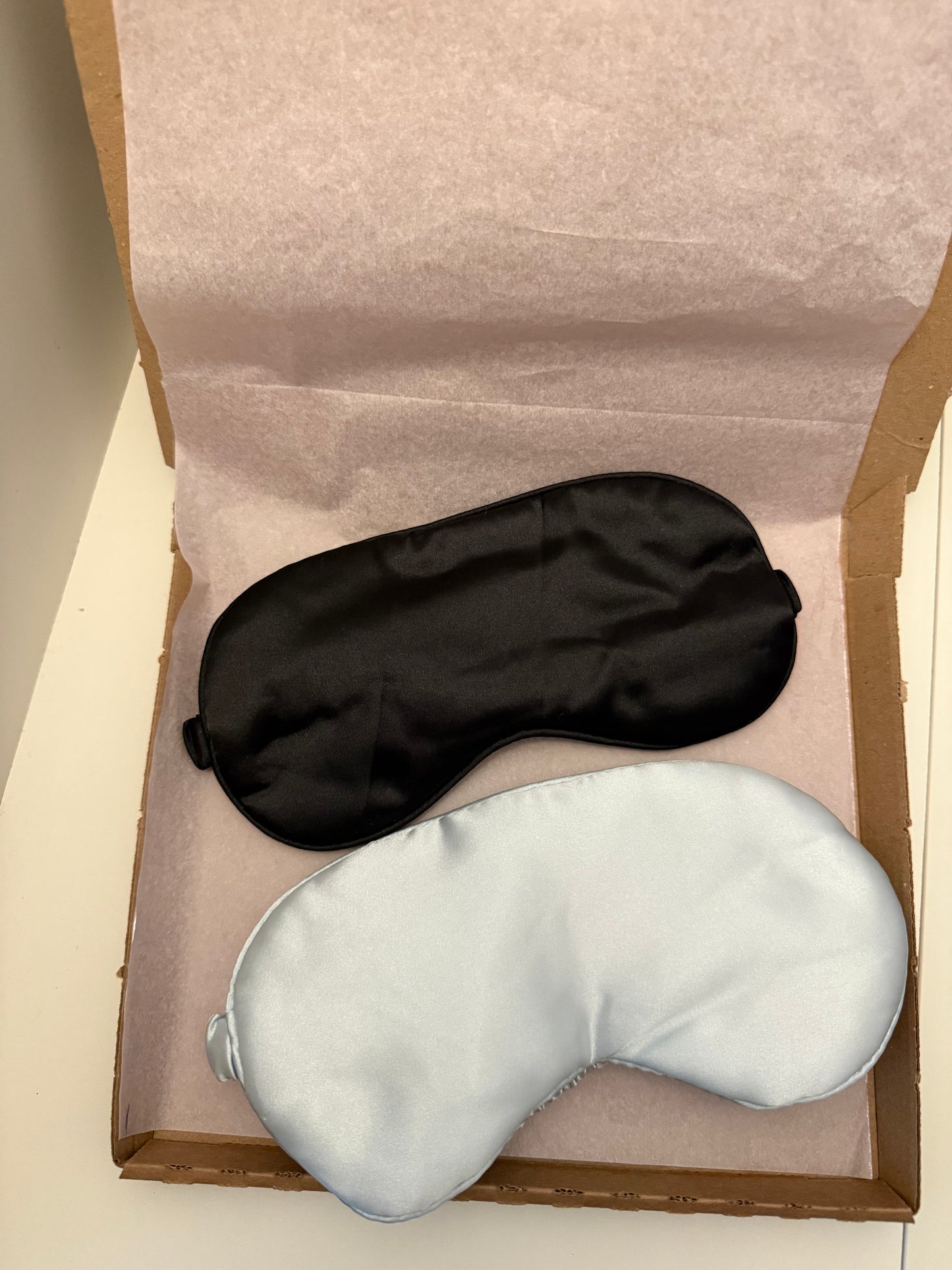 Two satin sleep masks, one black and one light silver, displayed inside an open brown cardboard box lined with light pink tissue paper.